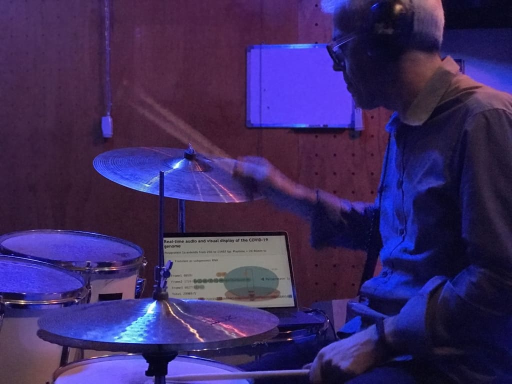 Dr Mark Temple drumming with a laptop that reads "Real-time audio and visual display of the COVID-19 genome" in background