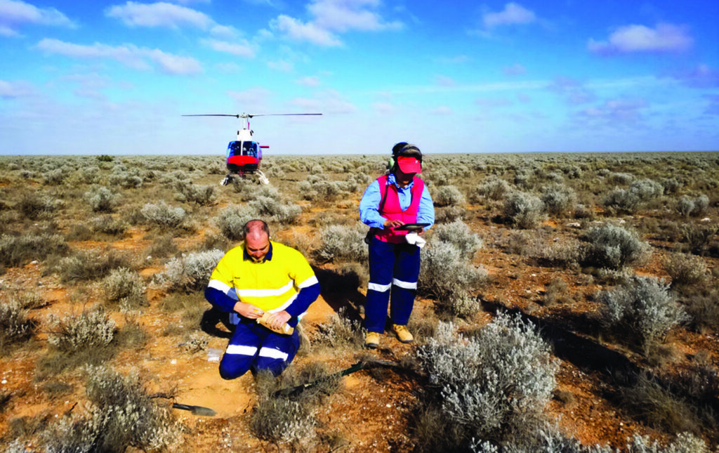 2 CSIRO staff taking geochemical samples and documenting them in the FAIMS mobile app during a GSSA/CSIRO sampling campaign in the Nullarbor Desert with helicopter in the background
