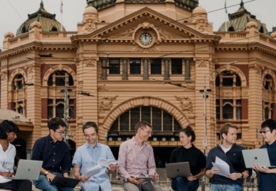 A group of people with laptops and documents sitting and conversing in front of the Edwardian Flinders Street railway station in Melbourne