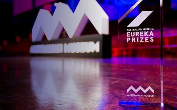 The crystalline Eureka Prize trophy set against a sculpture of the Australian Museum's logo in a room lit by purplish lights