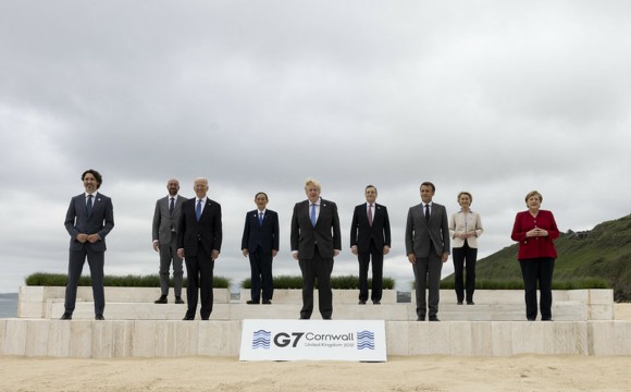 9 G7 leaders standing in 2 rows on a beach in business attire with a sign in front of them, reading "G7, Cornwall, United Kingdom, 2021"