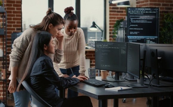 Three people gathering around a computer with code on the monitor, one of them typing