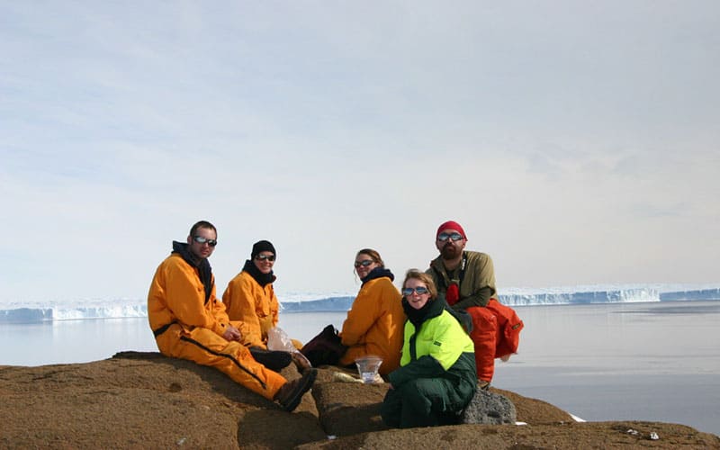 5 ACEAS students sitting on a rock with icebergs and ocean in the background, in Antarctica