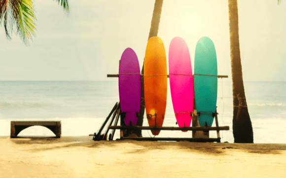 ARDC Holiday image of 4 surfboards in ARDC brand colours - purple yellow, pink and blue, on a beach with the sea in the background
