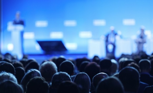 A conference hall full of people. A person is speaking at the lectern with two people sitting on the stage