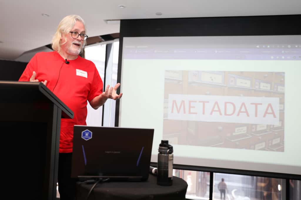 Tim Sherratt talking at the podium in front of a projection screen with  the word "metdata" on it