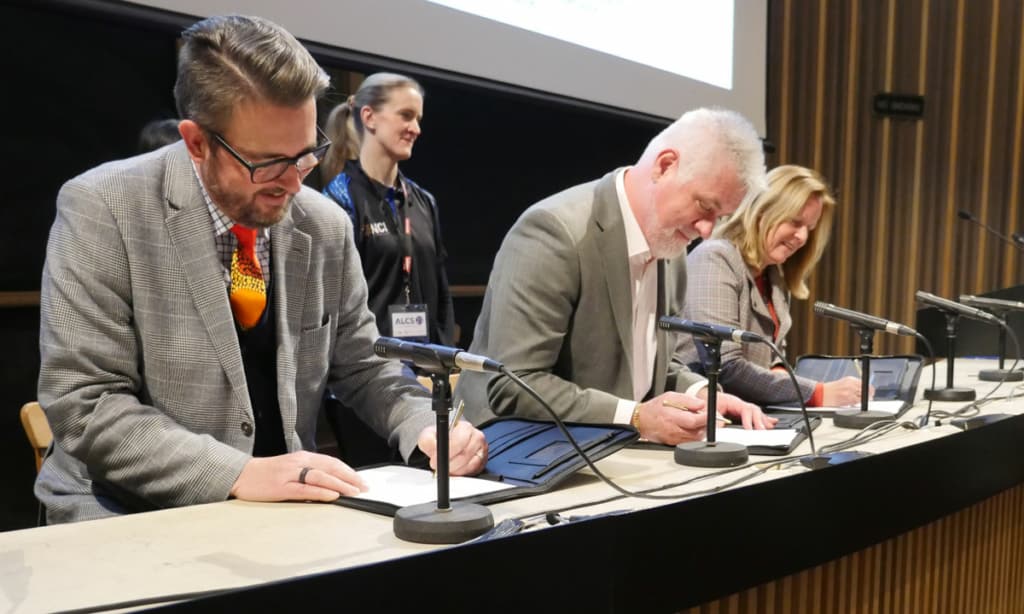 3 people signing a document on a table with a screen behind them.