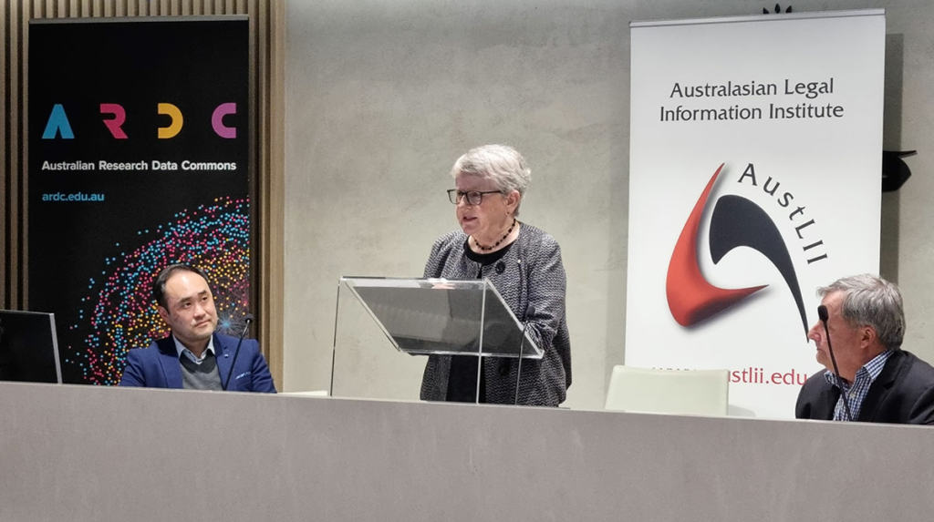The Hon Virginia Bell AC SC centre, at lecturn, speaking at the launch of the Australian Coronial Law Library. 2 people either side of her look on. 2 banners are up in the background.
