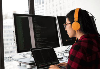 person with earphones on looking at 3 screens with code on them.