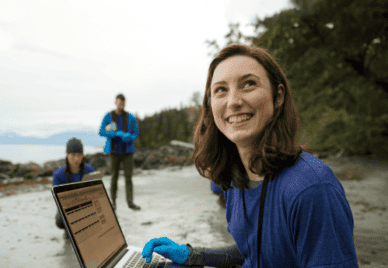 A researcher working with a laptop on a beach, smiling. Two other researchers in the background